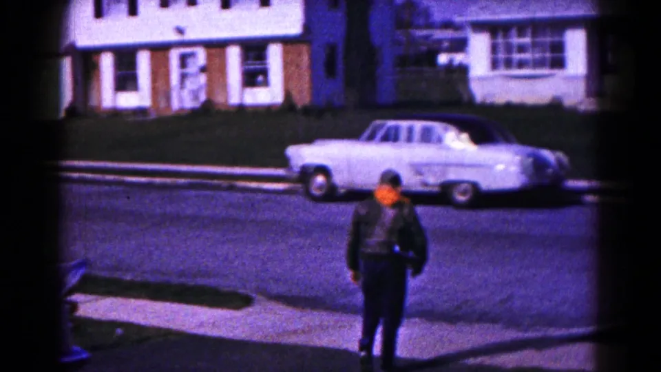 1961 Hagerstown sidewalk view with a walking person in a dark jacket and a white sedan parked along the residential street.