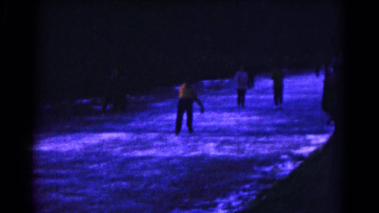 Several skaters move across a blue-lit outdoor ice surface at night.