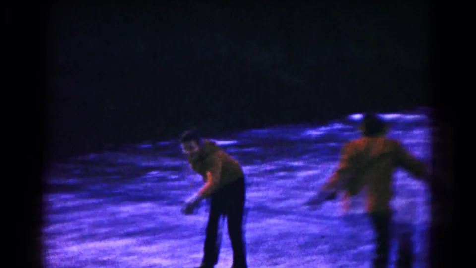 A close 8mm frame shows one skater bending slightly on the ice as another skater passes quickly at the right edge, with the frozen pond and dark background behind them.