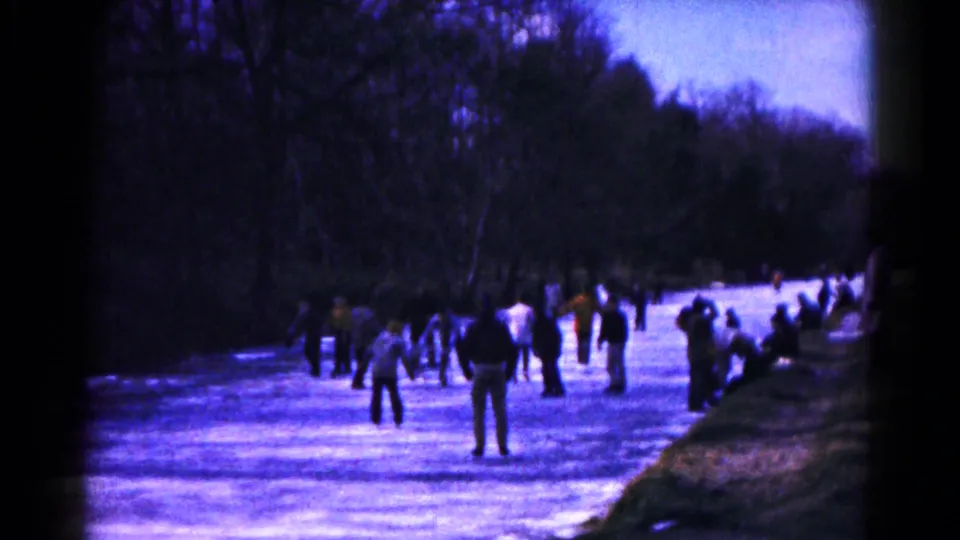 A wide home-movie frame from Hagerstown, Maryland shows people dispersed across a frozen pond, with some skating and others standing near the wooded shoreline.