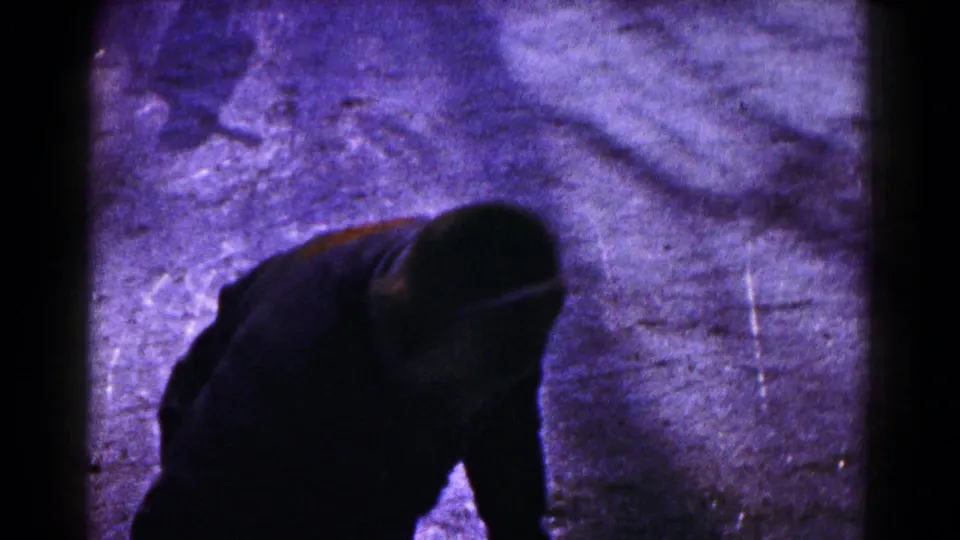 The home-movie camera catches a close, dark view of a skater bent forward while learning to balance on the ice.