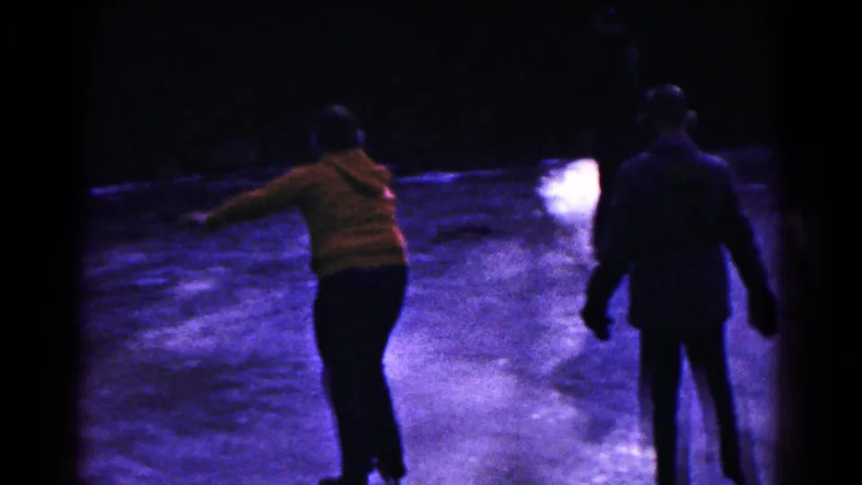 Two bundled figures skate away on the frozen lake, including one in an orange striped top, in 1961 Hagerstown footage.