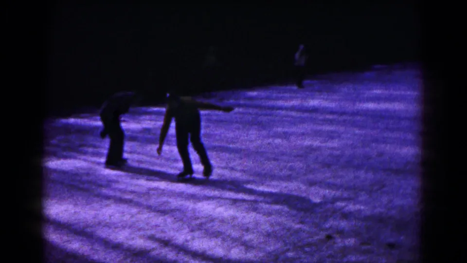 Home-movie frame of two skaters near each other on purple-lit ice, with another figure farther back.