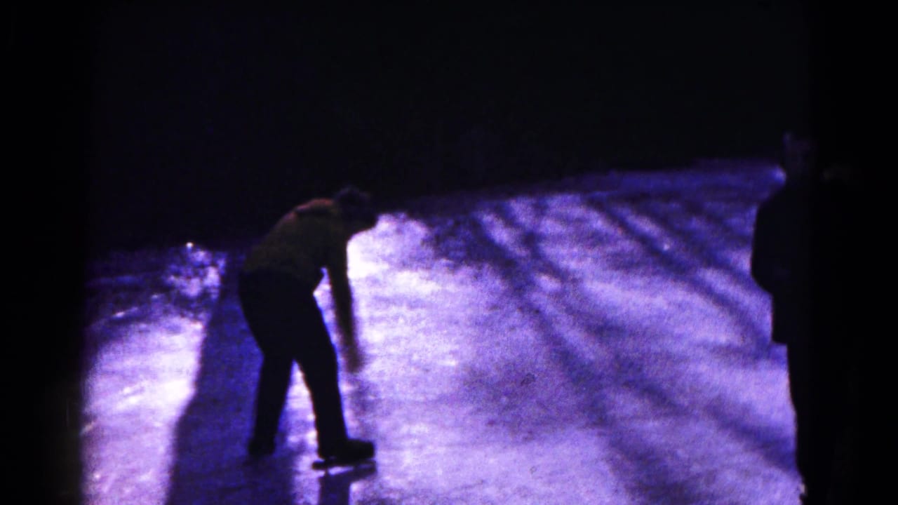 A skater leans forward while gliding on a dark, blue-lit frozen surface in 1961 Maryland home-movie footage.