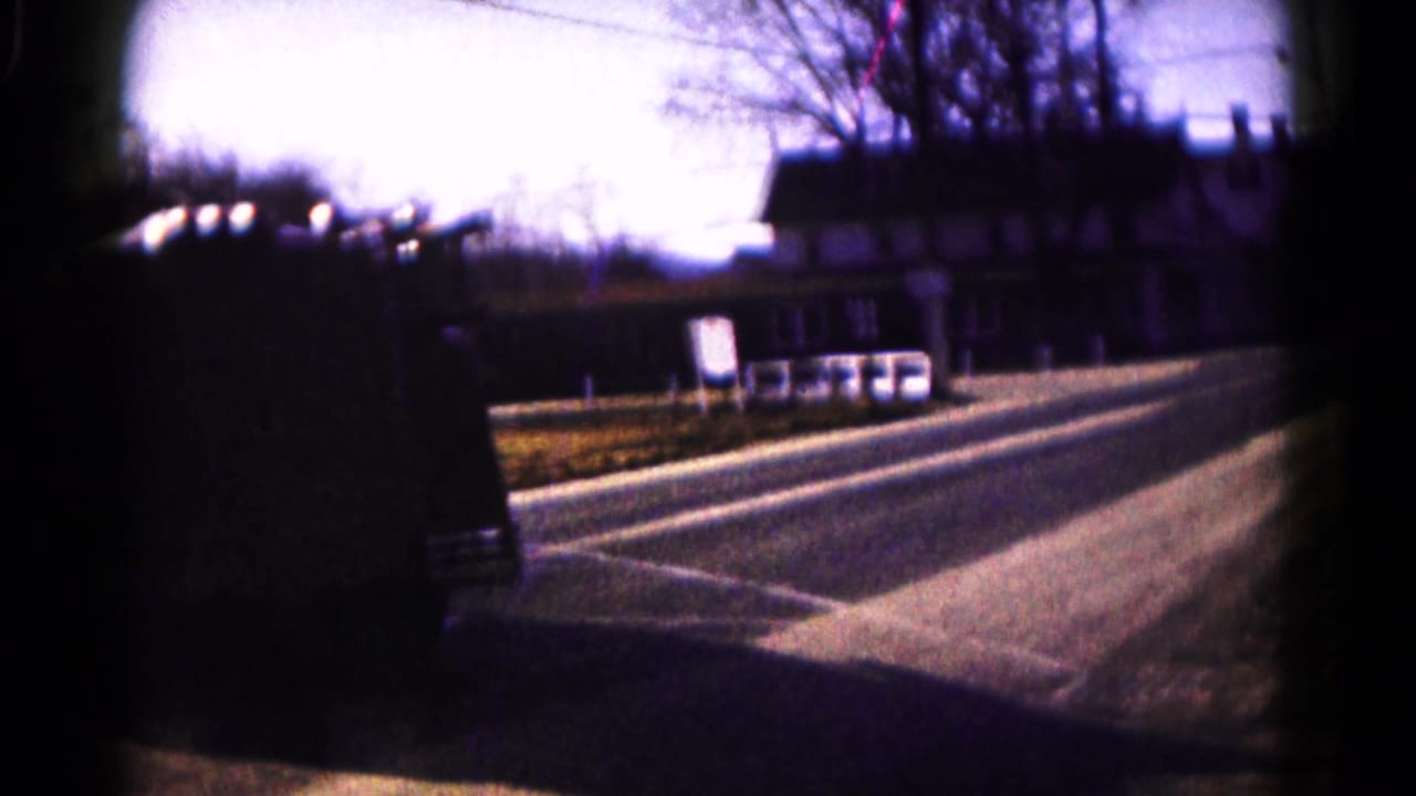 Quiet two-lane street with bare trees, roadside buildings, and a dark vintage car partly at the left edge.