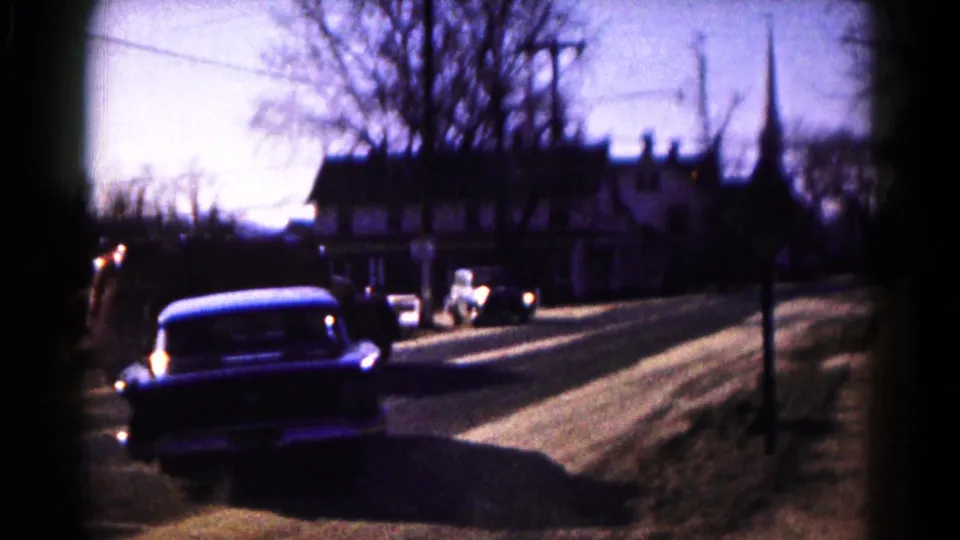 The blue sedan is seen from behind on a 1961 Hagerstown street, with another vehicle ahead and a church-like spire beyond the trees.