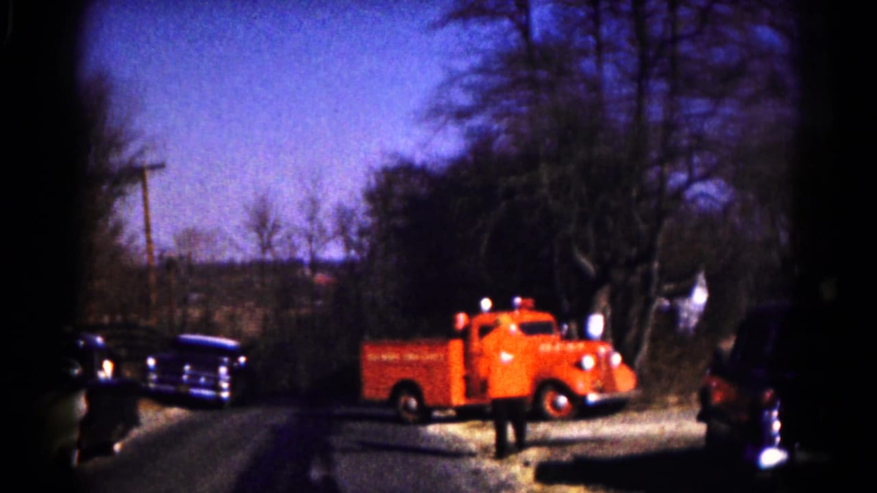 Red fire truck turning into a gravel driveway with dark vintage cars nearby.
