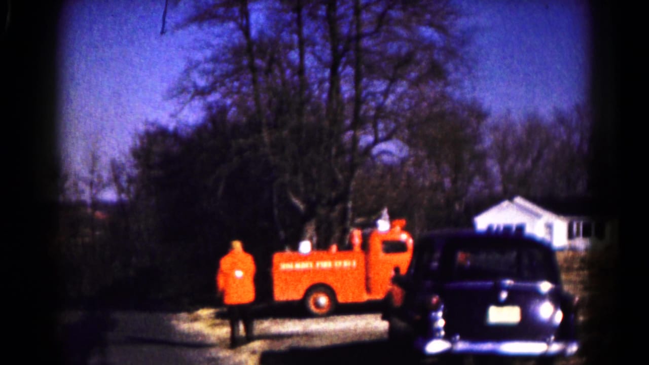 1961 Hagerstown frame with an orange-jacketed firefighter beside a red fire truck near a dark parked car and white house.