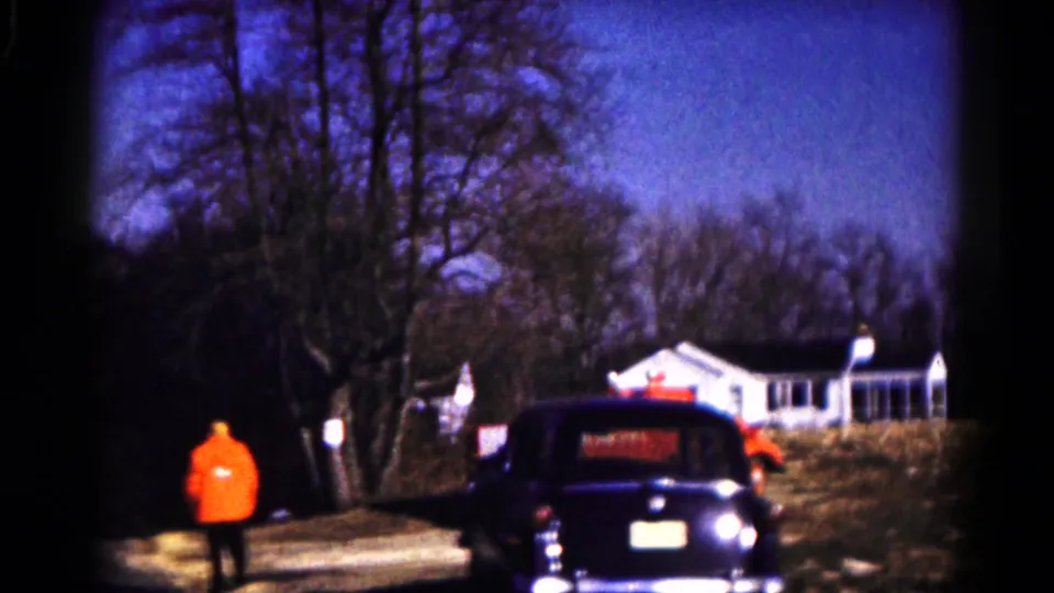 A home-movie view of a firefighter in orange standing near a driveway, with a dark vintage car and white house in view.