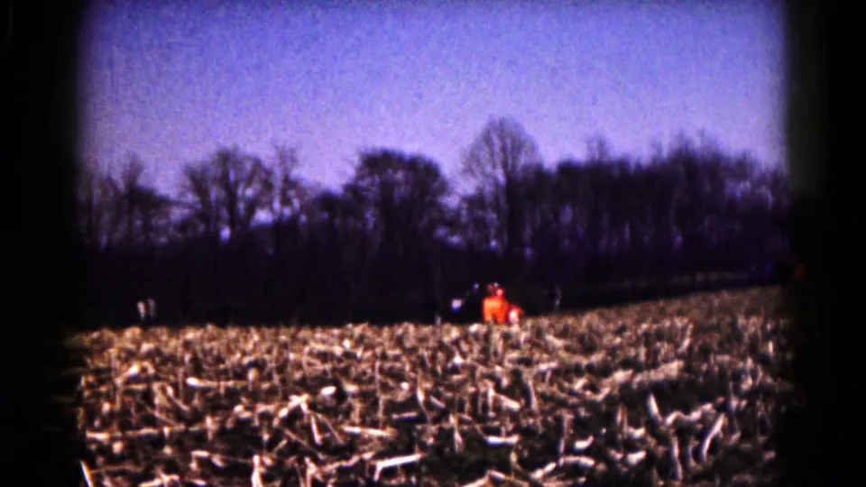 Home-movie view of a Hagerstown-area harvested field in 1961, with distant figures, a tractor, and leafless trees on the horizon.