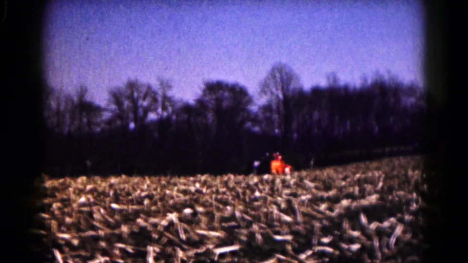 1961 Hagerstown-area 8mm frame showing a distant orange tractor crossing a harvested field below a dark treeline.