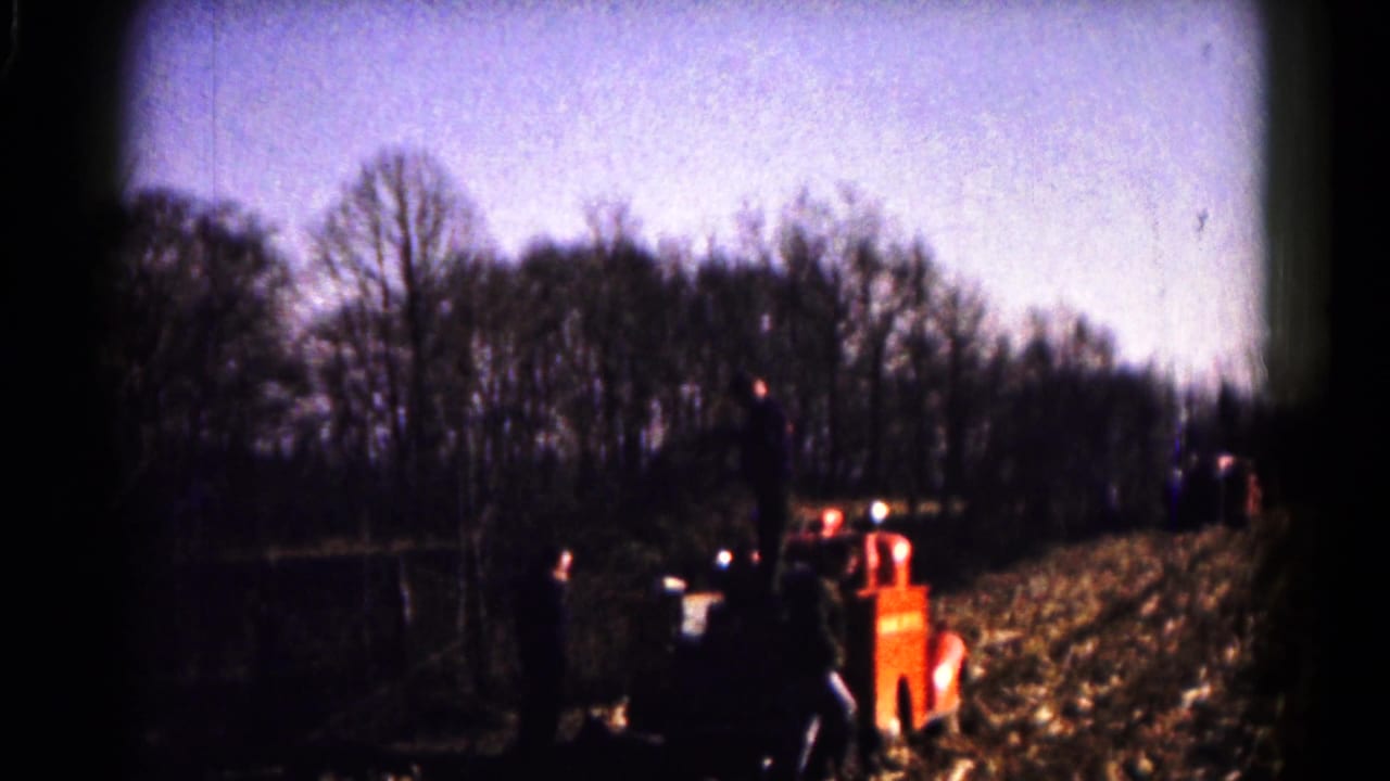 Distant view of the red fire truck in a rural field, with people nearby and a winterlike tree line behind it.