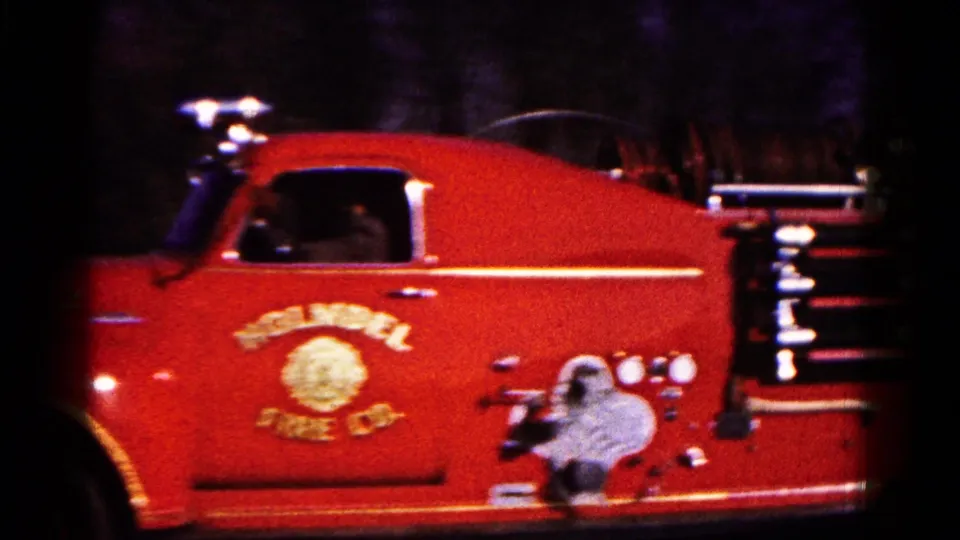 Close side view of the red fire truck with door lettering, a driver silhouette, mounted equipment, and dark 8mm frame edges.