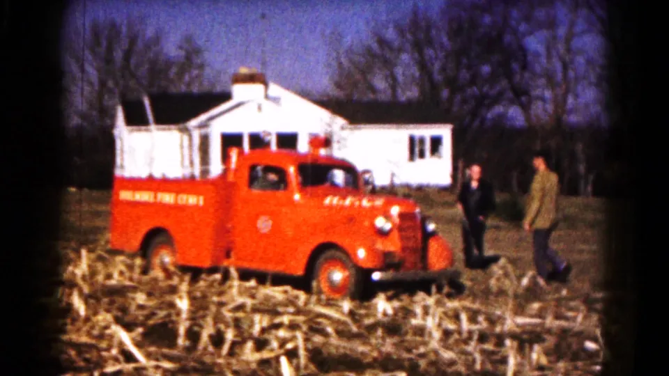 Two adults in period clothing stand near a red Hagerstown fire truck in a harvested field, with a white house behind them.