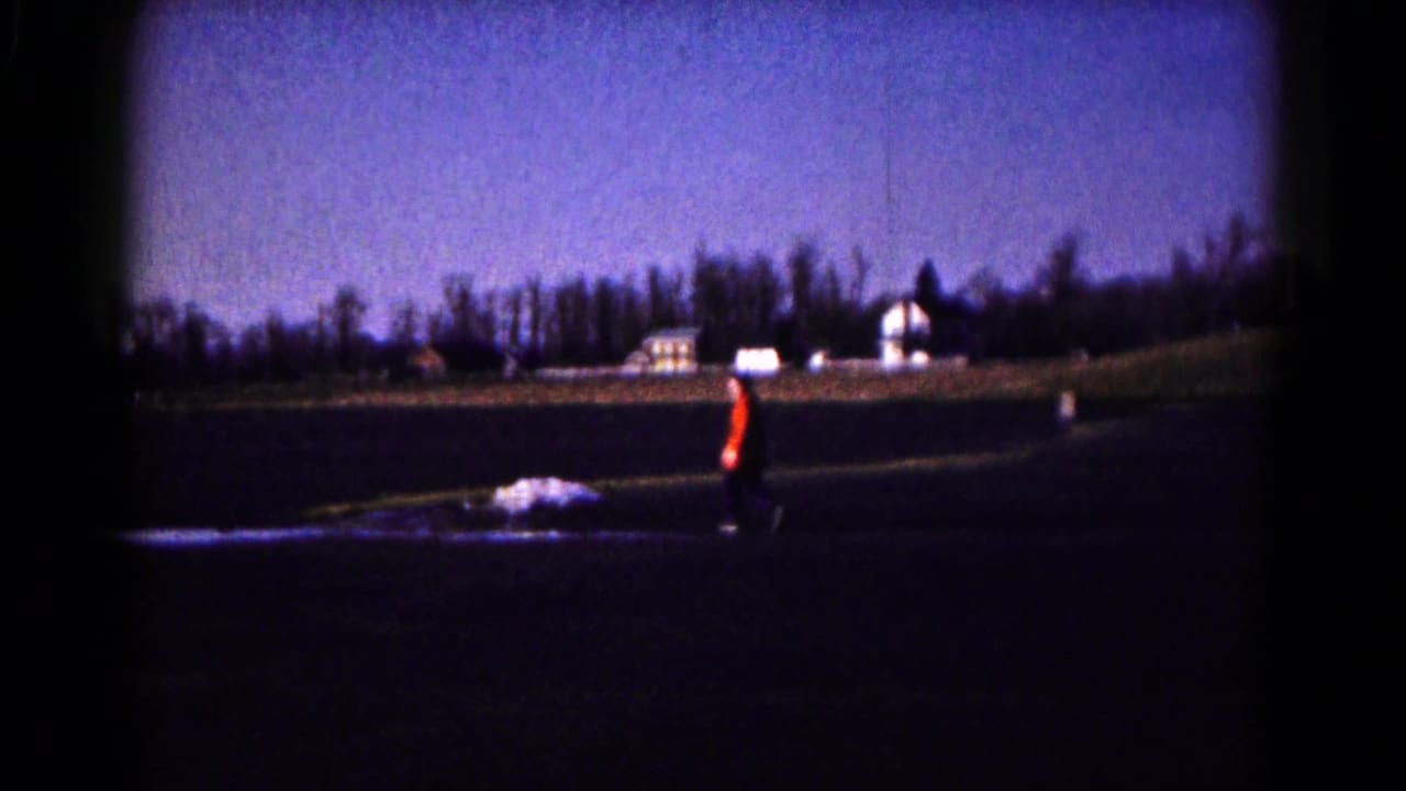 1961 Hagerstown 8mm frame with a distant person in an orange jacket walking across a field toward farm buildings.