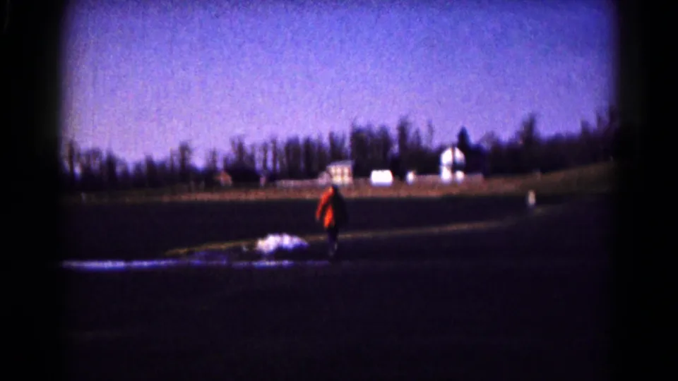 1961 Hagerstown home-movie frame showing a person in orange in a rural field below a line of distant houses and trees.