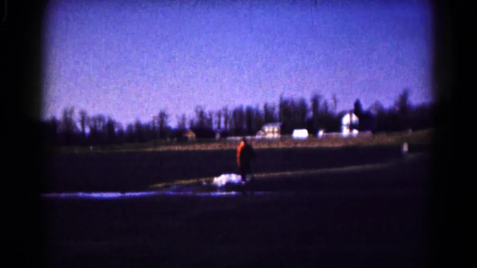 1961 Hagerstown 8mm frame of a lone person crossing a rural field, with distant farm buildings under a blue-purple sky.