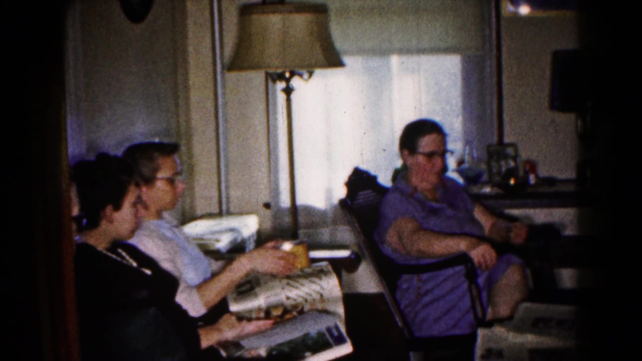 Three adults sit in a living room with newspapers, a snack bowl, chairs, lamps, and curtained windows.