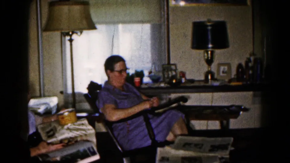 A woman in a purple dress sits in a living-room chair as another adult reads nearby with a small bowl on or near a printed page.