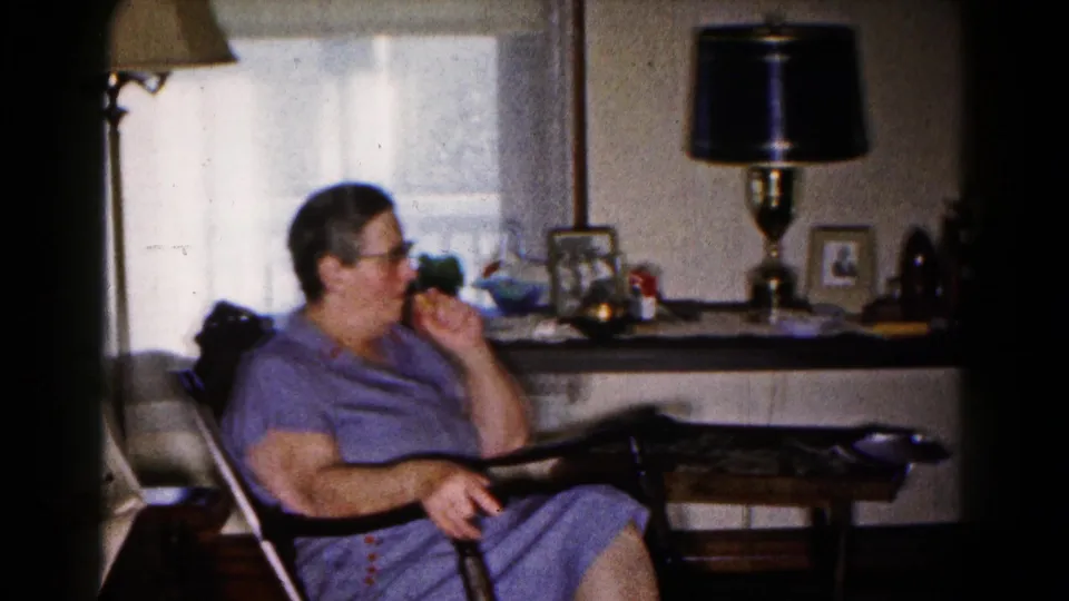 In the 1961 Hagerstown home interior, a woman in glasses and a purple dress sits near lamps, a side table, and curtained windows.