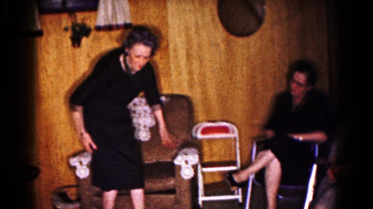 Woman in a black dress stands beside a chair while another woman sits nearby in a wood-paneled living room.