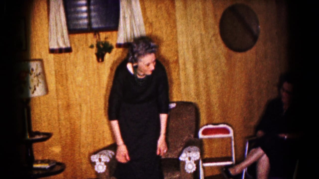 In the Hagerstown home interior, the woman in a black dress leans near the chairs while another seated woman appears at the frame edge.