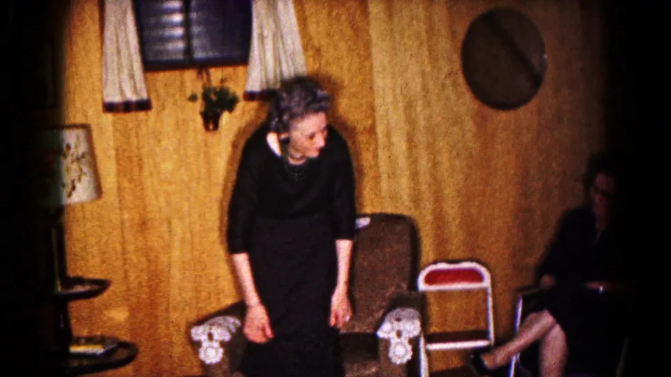In the Hagerstown home interior, the woman in a black dress leans near the chairs while another seated woman appears at the frame edge.