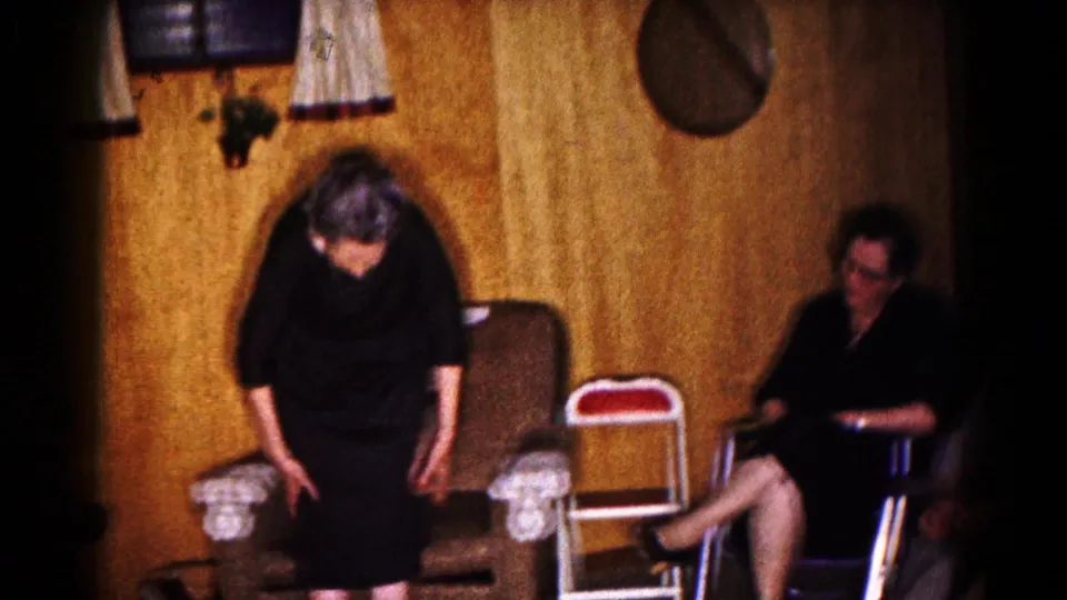 The 1961 home-movie frame shows the woman leaning forward near an upholstered chair, with a seated companion partly visible in the living room.