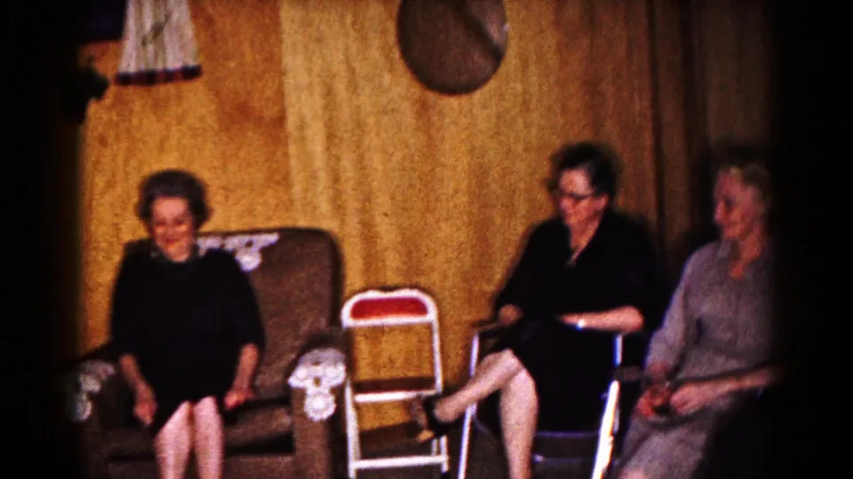 Three adult women sit in a 1961 Hagerstown living room, with folding chairs, an upholstered chair, curtains, and wall decor visible behind them.