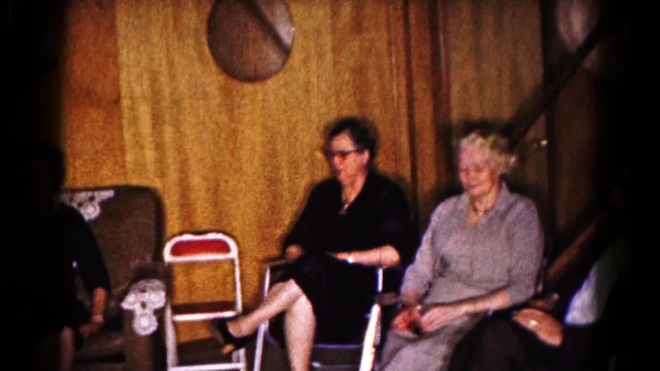 1961 Hagerstown home-movie frame with adults seated in a paneled interior, including two women and a partial figure at left.