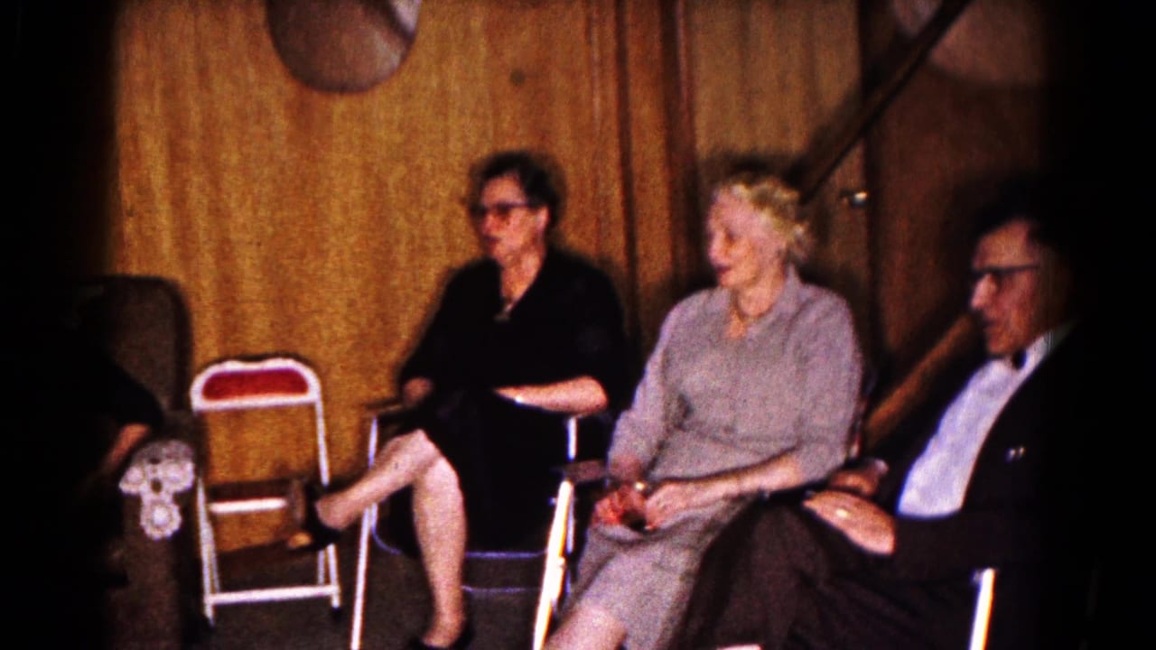 Two women and a suited man sit beside a folding chair in a paneled interior.