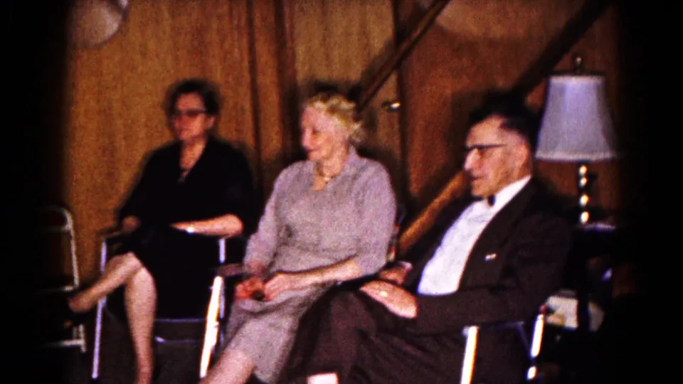 1961 Hagerstown home-movie frame with two women and a suited man seated in a warm paneled room near a table lamp.