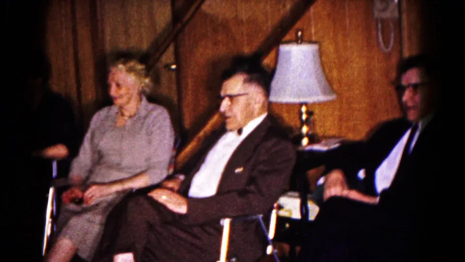 1961 Hagerstown frame showing seated adults in a paneled room, with a table lamp, stair rail, and formal mid-century clothing visible.