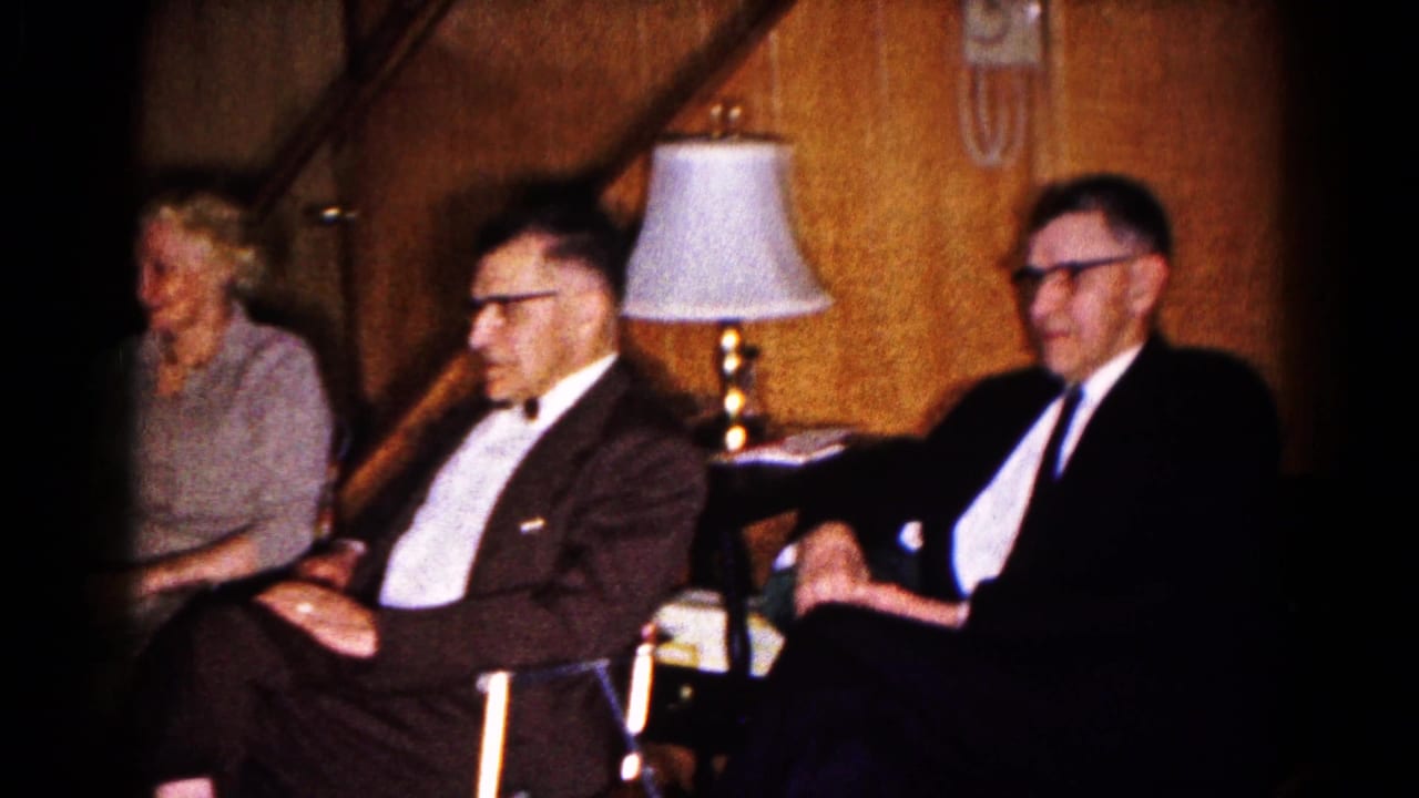 Three formally dressed adults seated in a wood-paneled room beside a lamp and staircase.