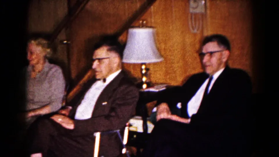 In a 1961 Hagerstown home-movie frame, three adults sit formally in a wood-paneled room near a table lamp and staircase.