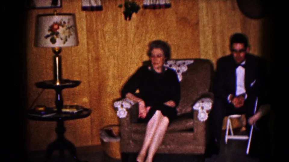 A formally dressed woman and man sit in a Hagerstown interior with patterned armchair covers, a lamp, and framed wall decoration.
