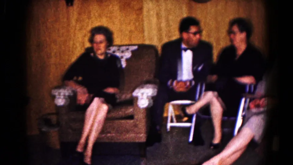 Several adults in formal clothing appear seated and conversing in a 1961 Hagerstown wood-paneled room with upholstered seating.