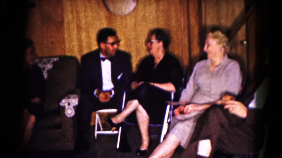 Four adults sit close together in a Hagerstown interior, with a man in formalwear and women in dresses near a staircase and armchair.