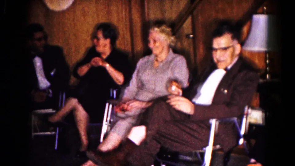 In 1961 Hagerstown home-movie footage, older adults sit in conversation beside wood-paneled walls and a table lamp.