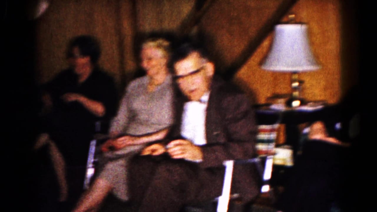 Older adults sit near a table lamp, with one man looking down at his hands.