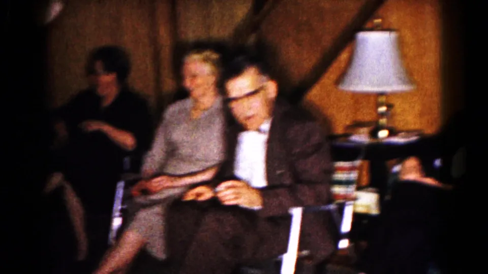 Older adults at a 1961 indoor gathering in Hagerstown sit near a lit table lamp and wood-paneled wall.