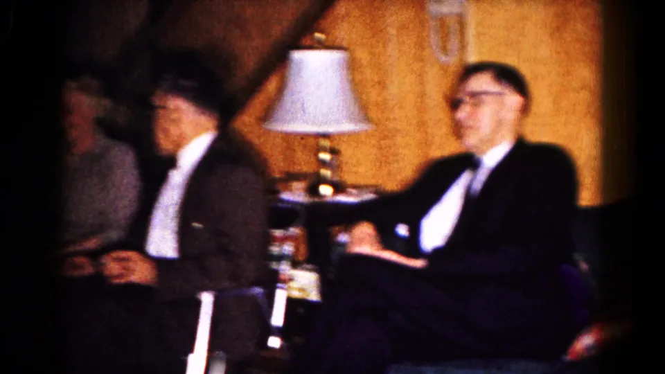 A formally dressed man sits beside a lamp while other party guests appear blurred in the 1961 Hagerstown interior.