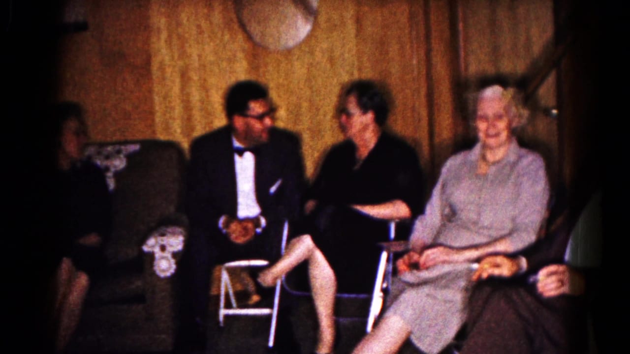 Adults seated in a wood-paneled living room, including a man in a tuxedo and women in dresses.