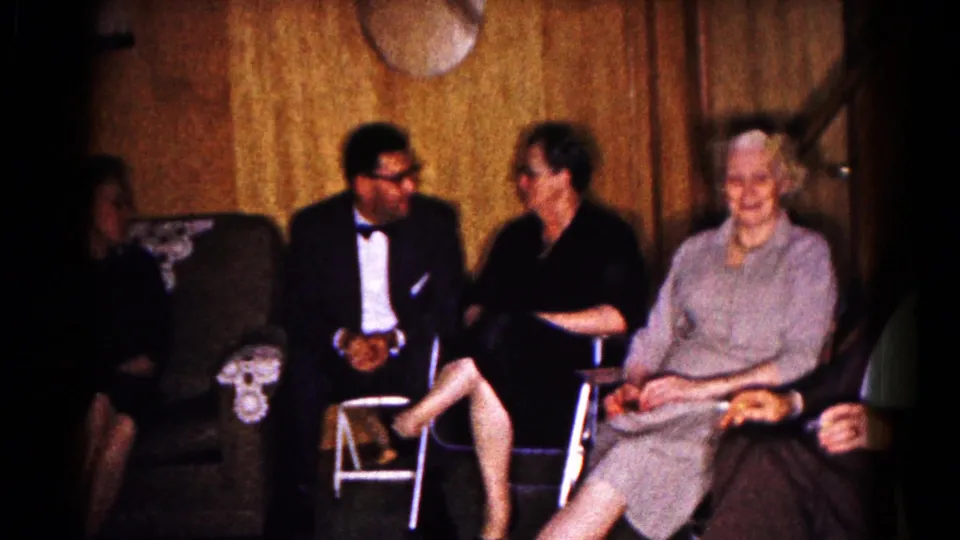 1961 Hagerstown home-movie frame showing adults seated in a wood-paneled living room during a family gathering.
