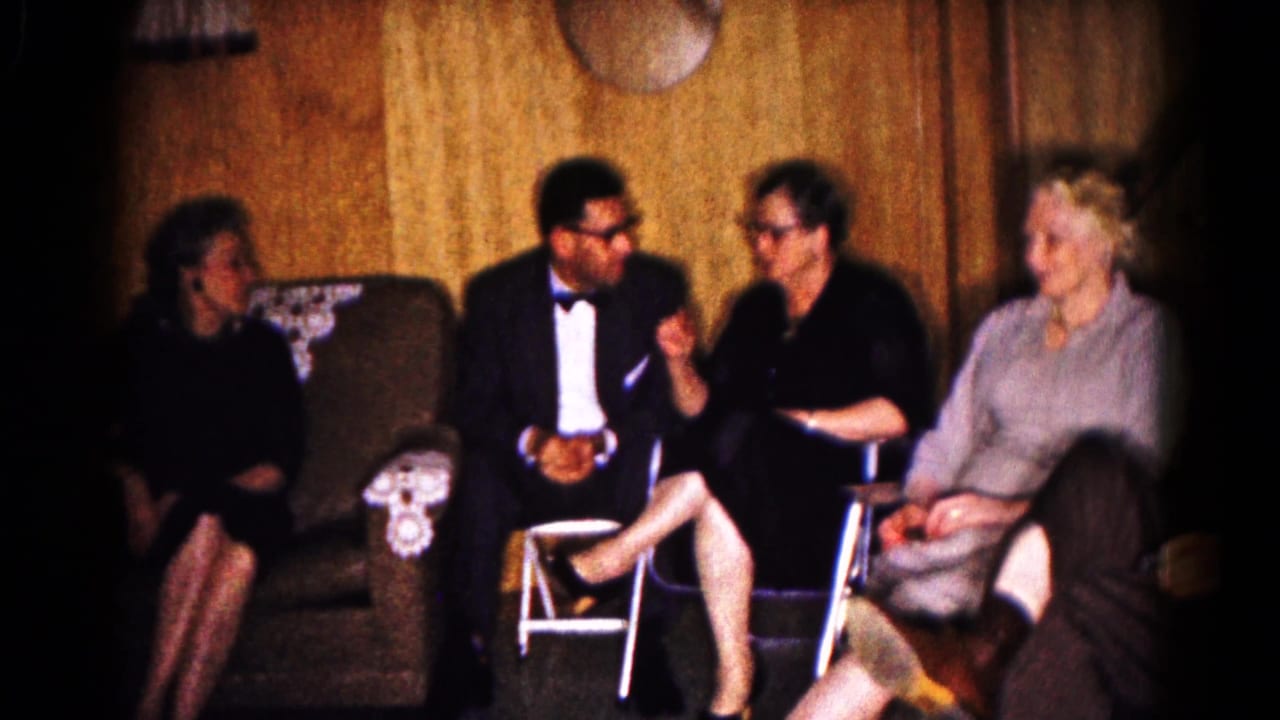 Seated adults talk and smile in a living room with wood paneling and upholstered chairs.