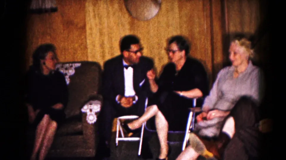 Adults in formal and semi-formal clothing converse in a 1961 Hagerstown living room home-movie scene.