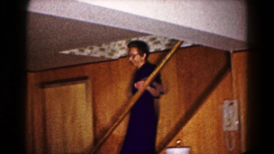 Woman in a dark dress stands by a staircase with a long pole in a 1961 Hagerstown home interior.