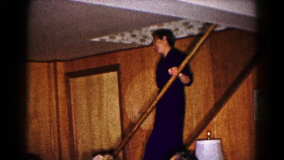A woman in a dark dress pauses beside the stairs with a long pole in a 1961 Hagerstown home-movie frame.