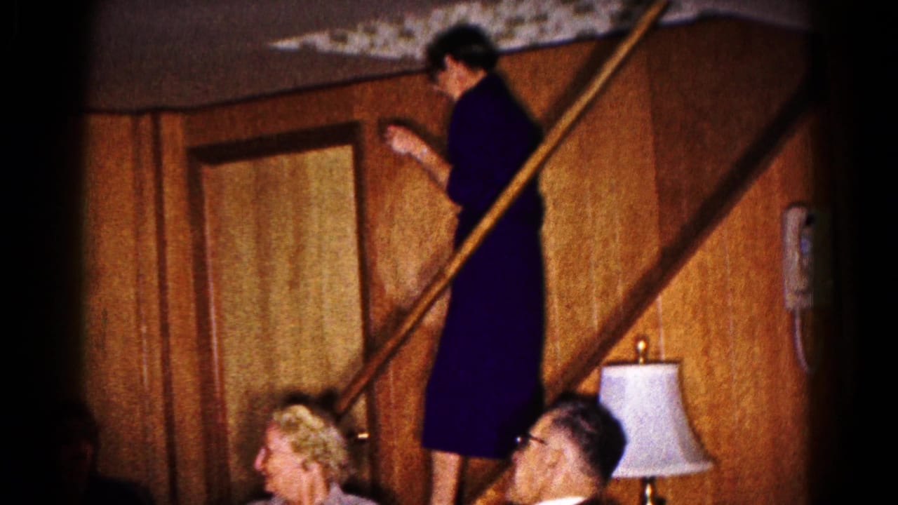 1961 Hagerstown home-movie frame showing a woman on a wooden staircase addressing adults seated below in a paneled living room.