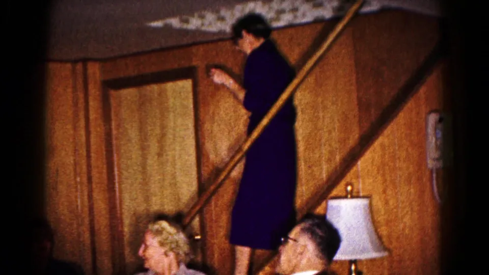 1961 Hagerstown home-movie frame showing a woman on a wooden staircase addressing adults seated below in a paneled living room.